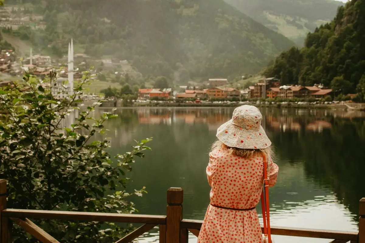 A woman in a sundress and hat stands by a lake, enjoying the scenic mountain town view.