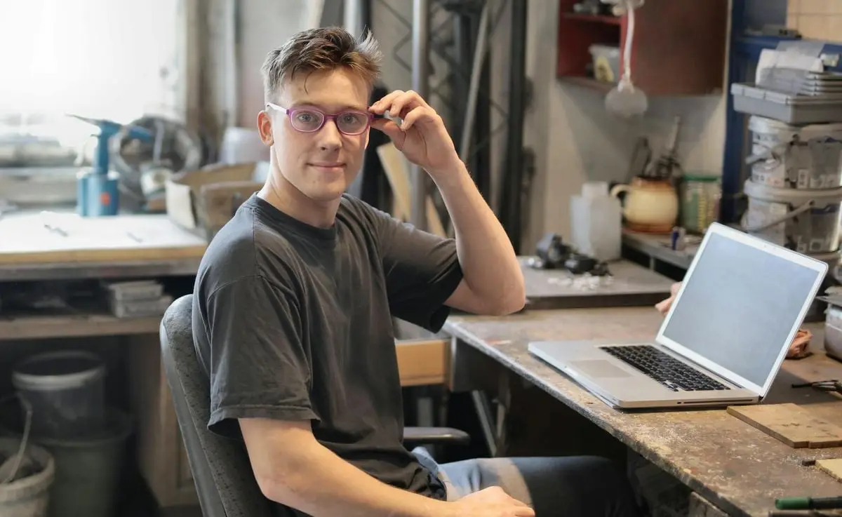 Young man in glasses working remotely on laptop in a workshop setting.