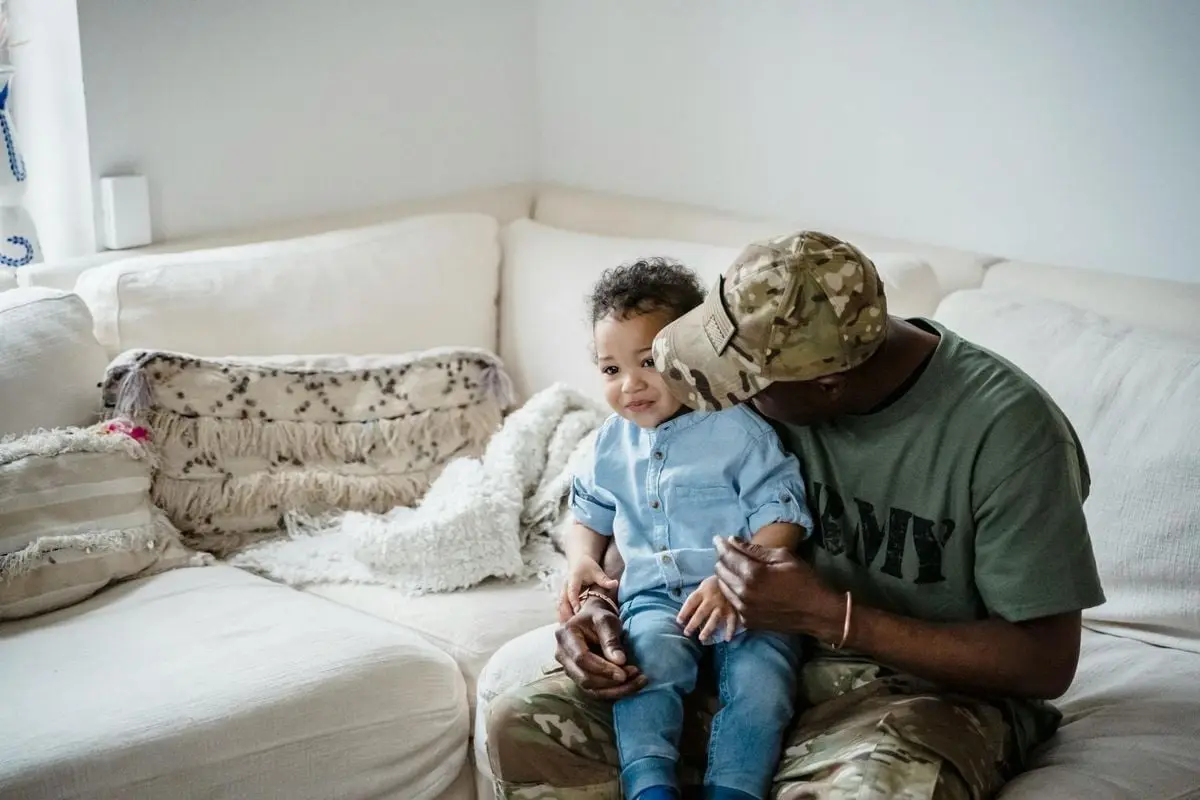 A loving father in army attire embraces his child at home on a cozy couch.