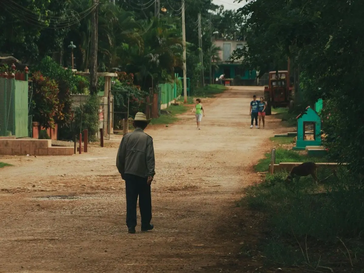 A man in a hat walks down a rural dirt road in a small town setting on a warm day.