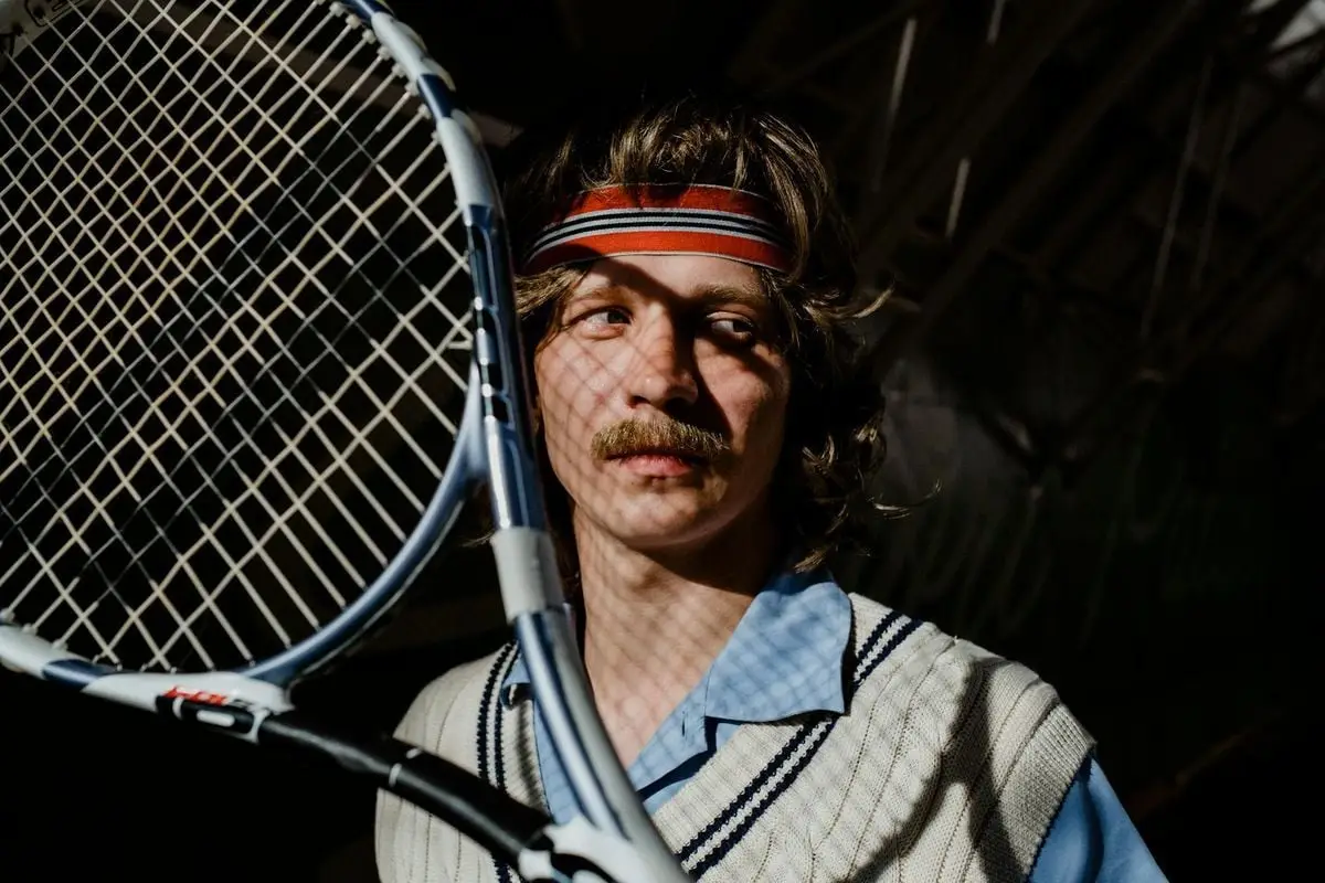 Vintage-style portrait of a man with a mustache holding a tennis racket indoors.