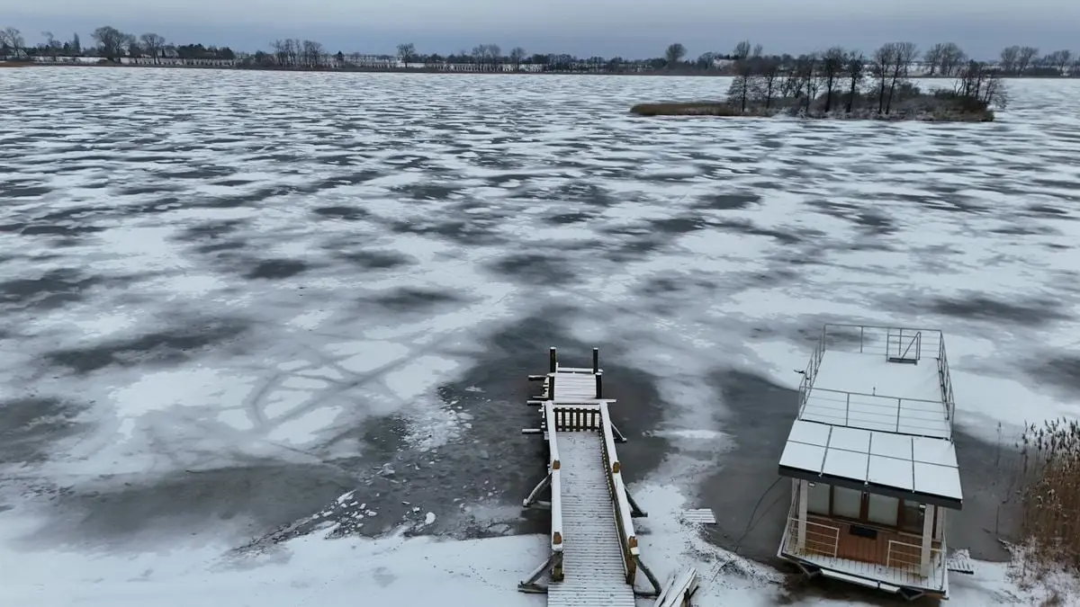 A scenic view of a frozen lake with a snow-covered pier and a small docked house in winter.