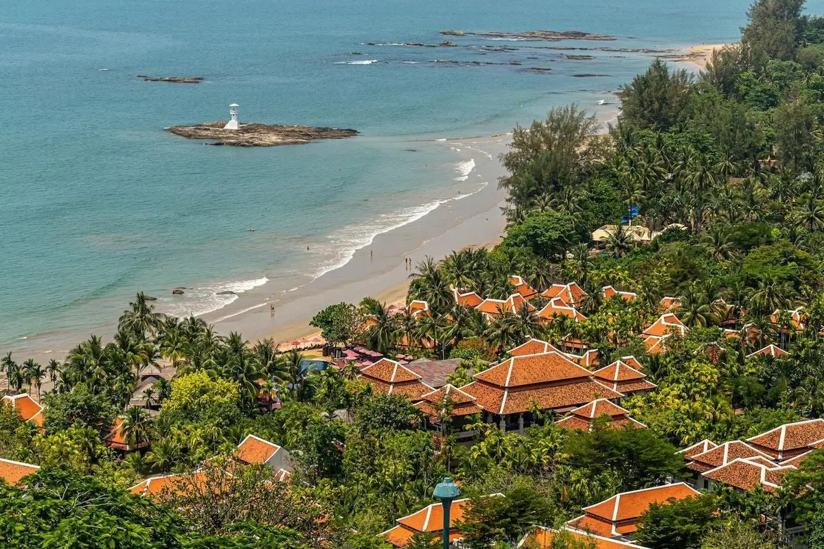 A picturesque beachfront village with terracotta roofs, palm trees, and a distant lighthouse.