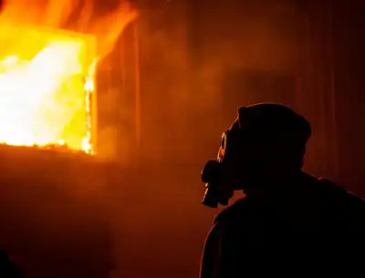 Silhouette of a person wearing a gas mask in front of a burning building at night.