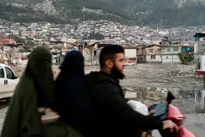 People on a scooter in Antakya, Türkiye, with a mountainous town backdrop, reflecting daily life and culture.