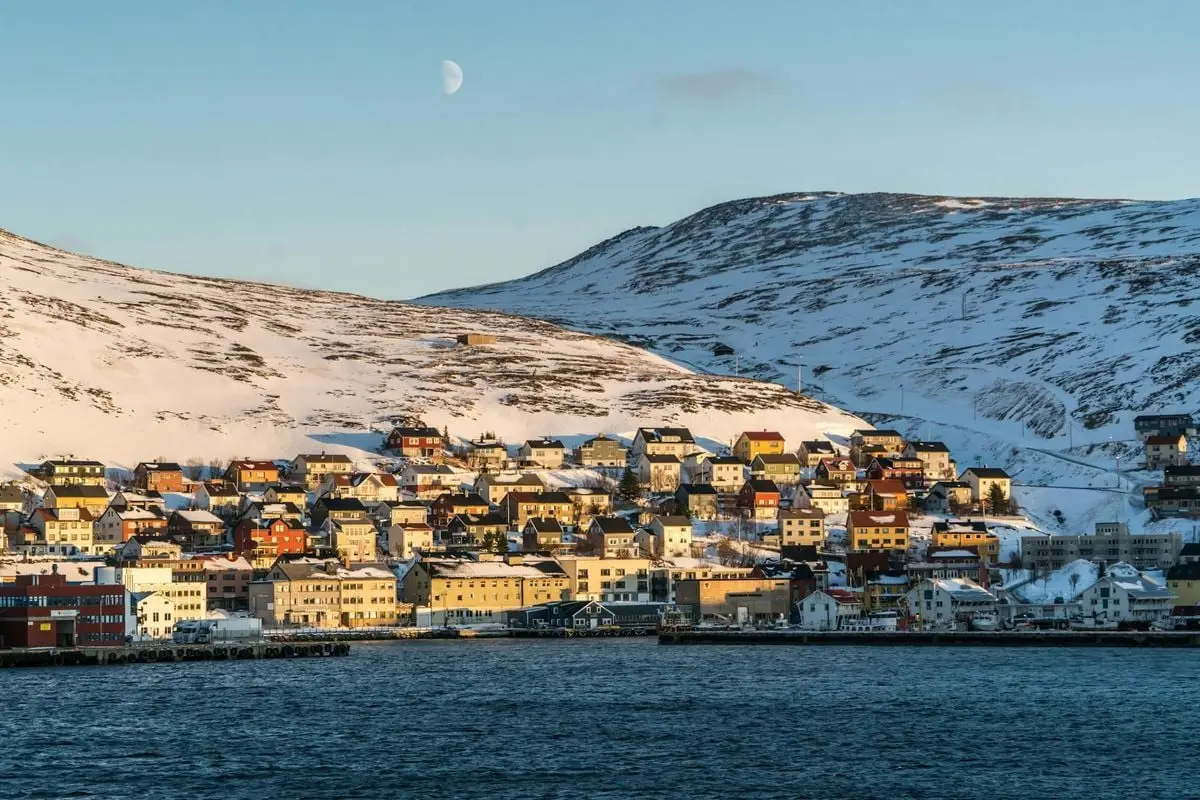 Charming Norwegian town with colorful houses on snowy hills by the sea under a bright moon.