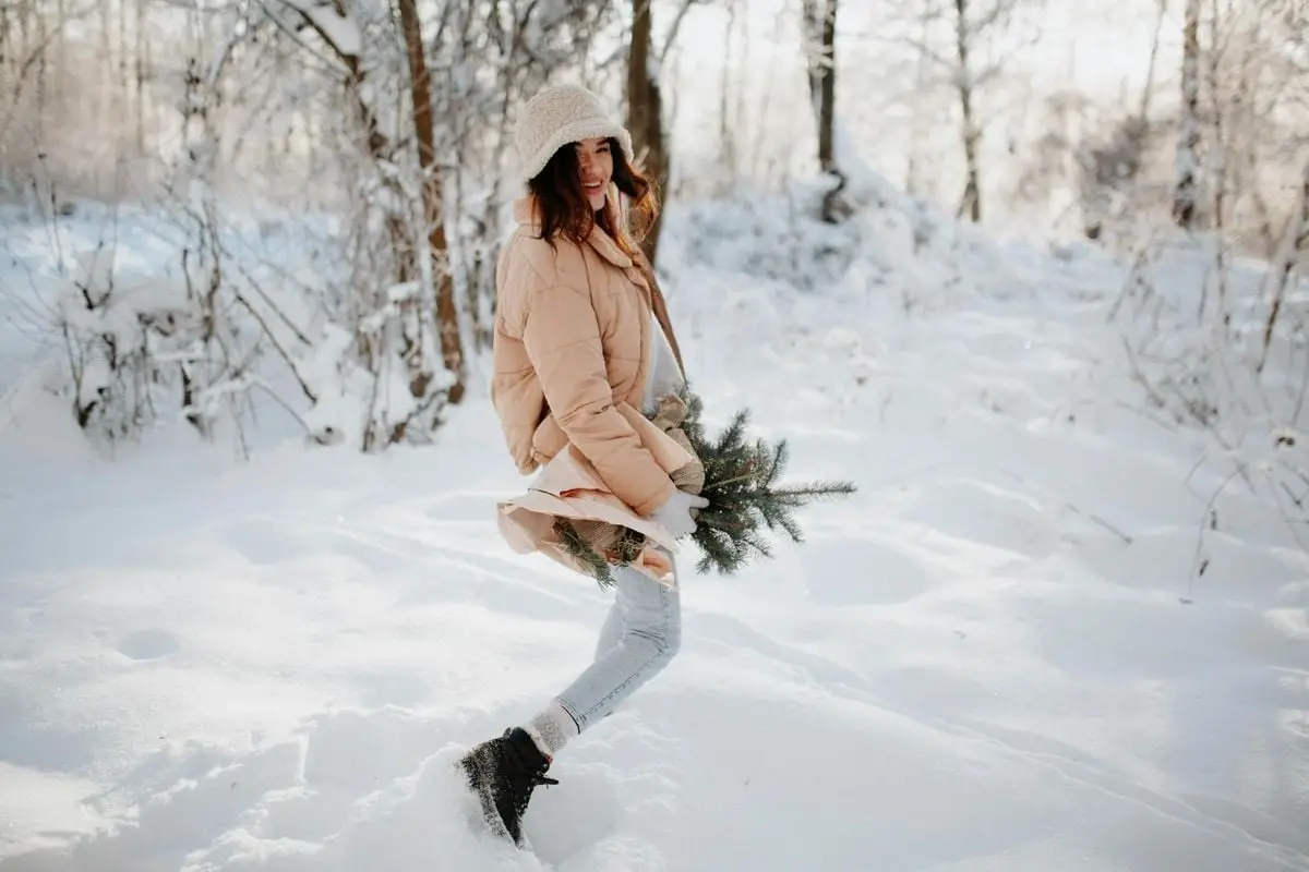 Smiling woman in winter attire holding fir branches, enjoying snow-covered forest.