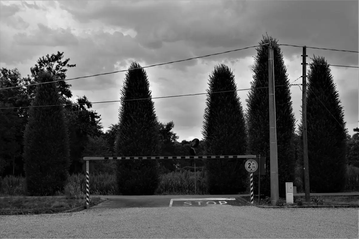 Black and white photo of a rural parking lot entrance with trees and cloudy sky.
