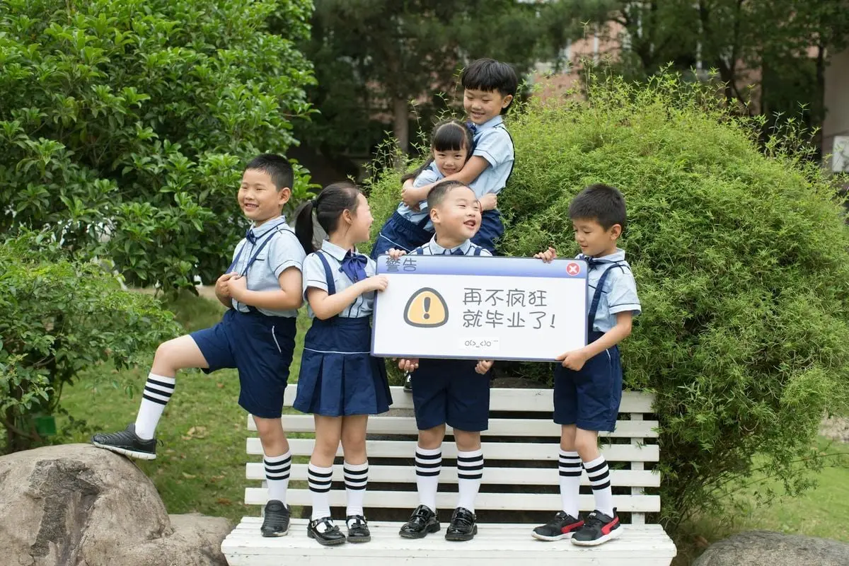 Children in school uniforms having fun outdoors, holding a playful sign on a bench.
