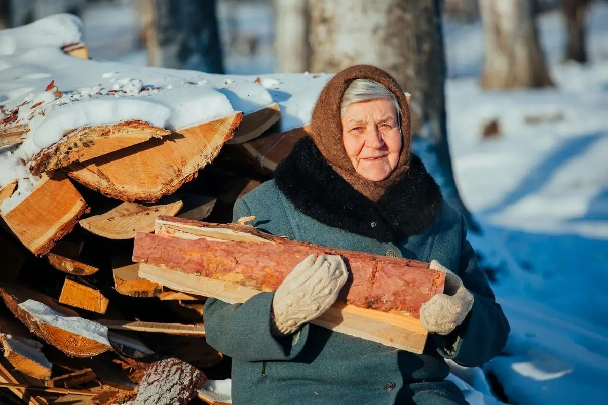 Elderly woman holding firewood in snowy winter landscape, wearing warm clothes.