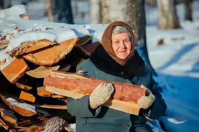 Elderly woman holding firewood in snowy winter landscape, wearing warm clothes.