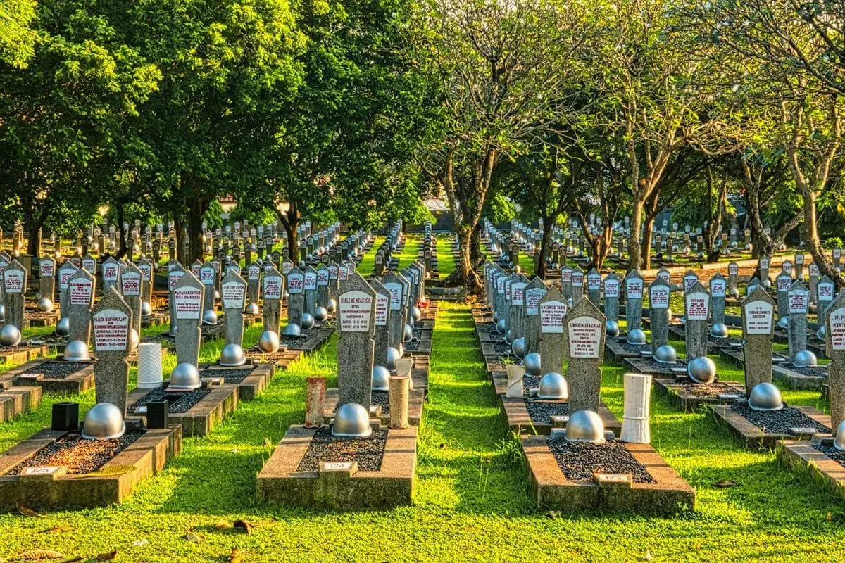 Rows of tombstones with military helmets located on grassy main heroes cemetery with tall lush trees and bright sunlight in Kalibata
