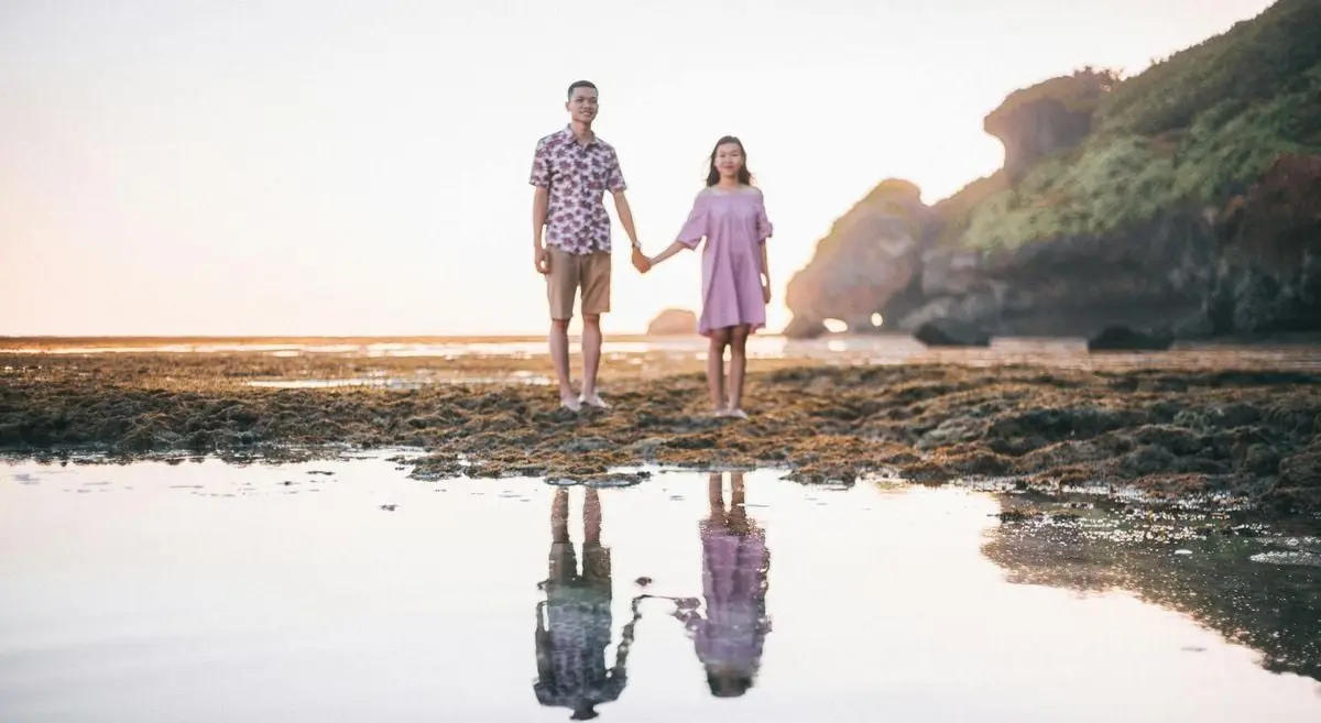 A young couple holding hands on a beach with their reflection in water under a serene sky.