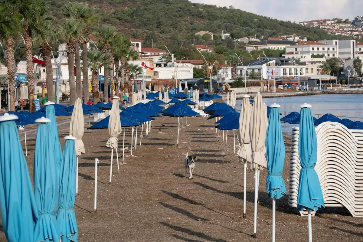 Empty beach in Foça, İzmir with blue umbrellas against a quaint town backdrop.