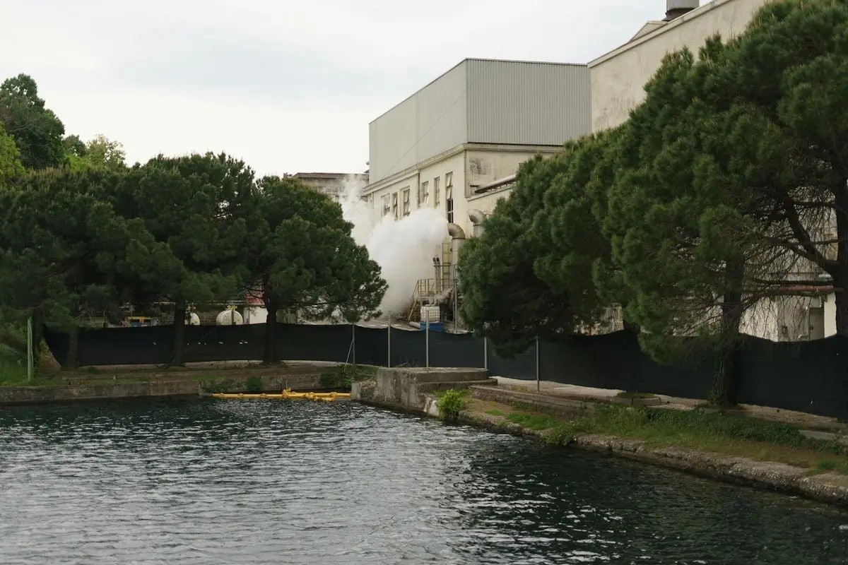 View of an industrial building with steam by a lakeside, surrounded by trees in Toscolano Maderno, Italy.