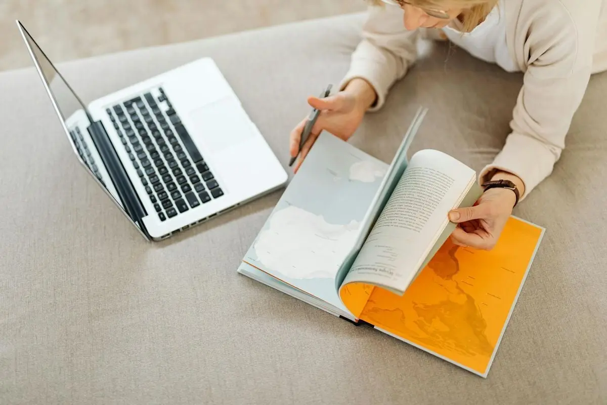 Overhead view of a woman using laptop and opening a map book for research.