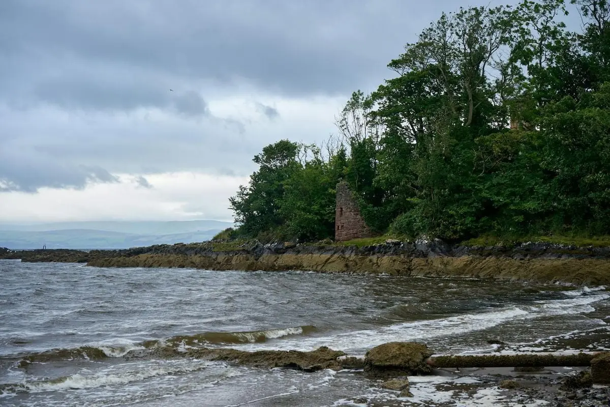 Dramatic coastline with waves, trees, and historical ruins under a cloudy sky.