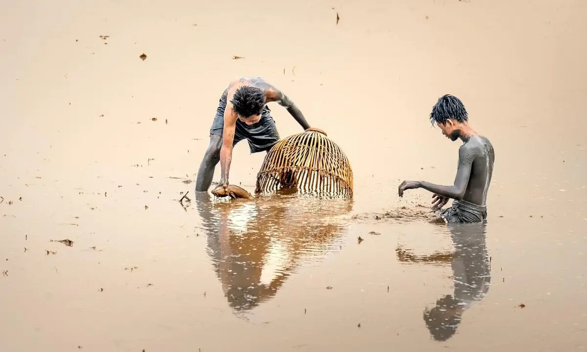 Two young men fishing in a muddy wetland using bamboo cages, showcasing rural life and traditional methods.