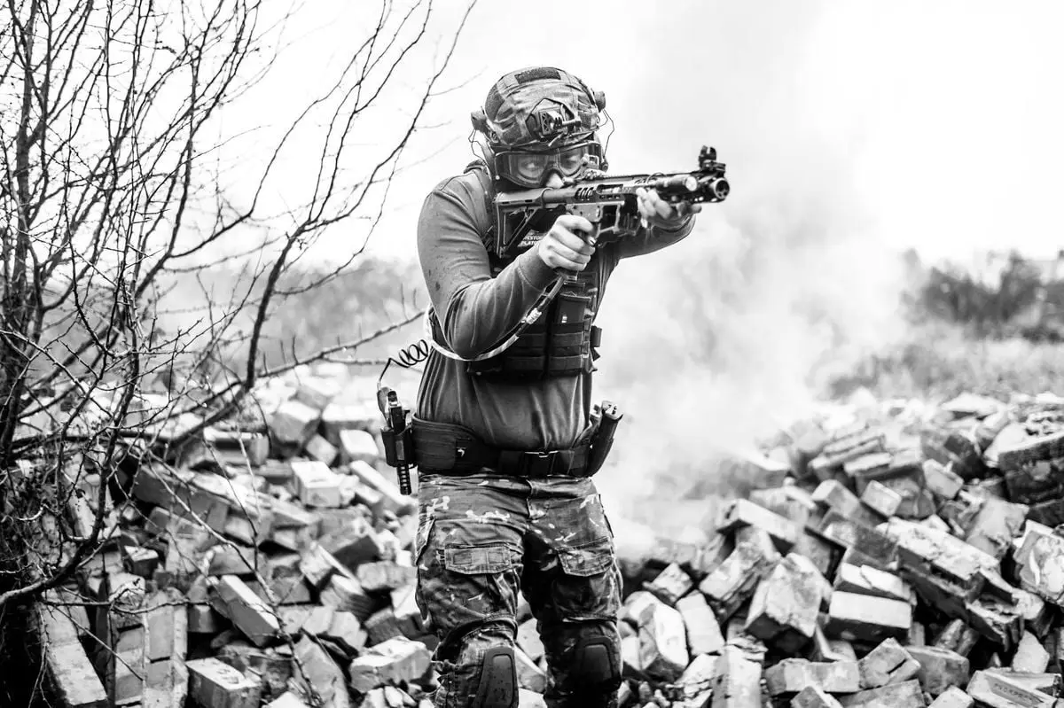 A soldier in full gear aims his rifle during a tactical training session on a rubble-strewn field.