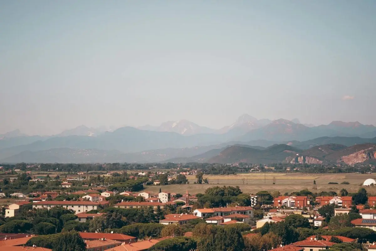 Aerial view of Pisa, Italy, highlighting the Apuan Alps in the background during a warm summer day.
