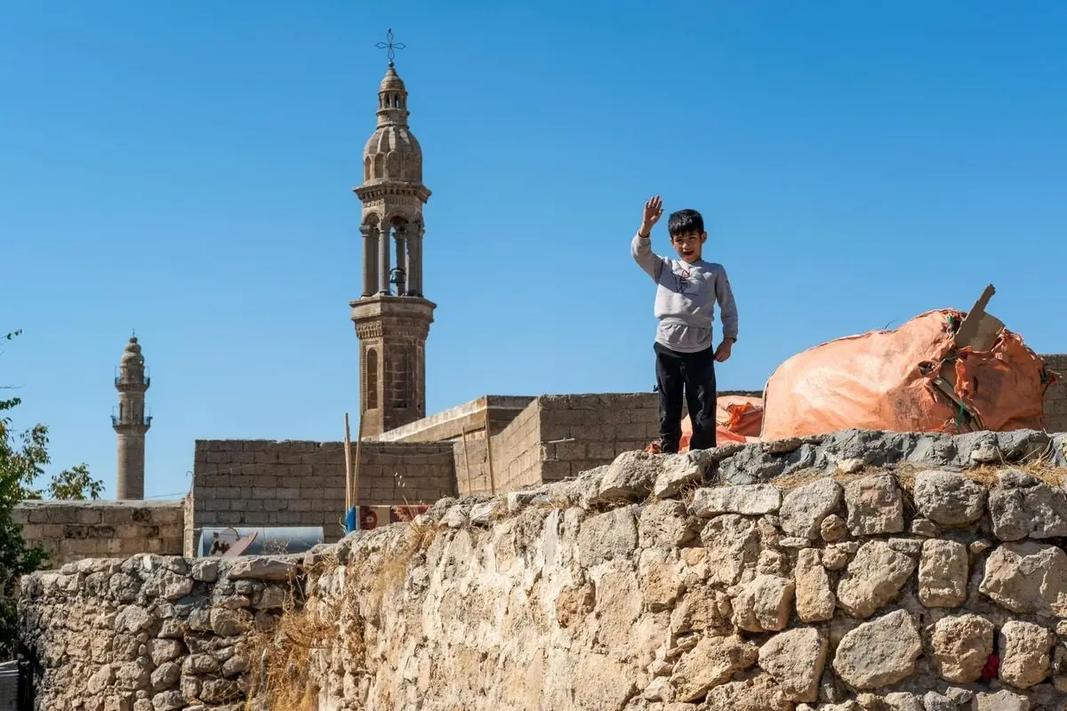 A smiling boy waves from a stone wall in Midyat, with historic bell towers in the background.