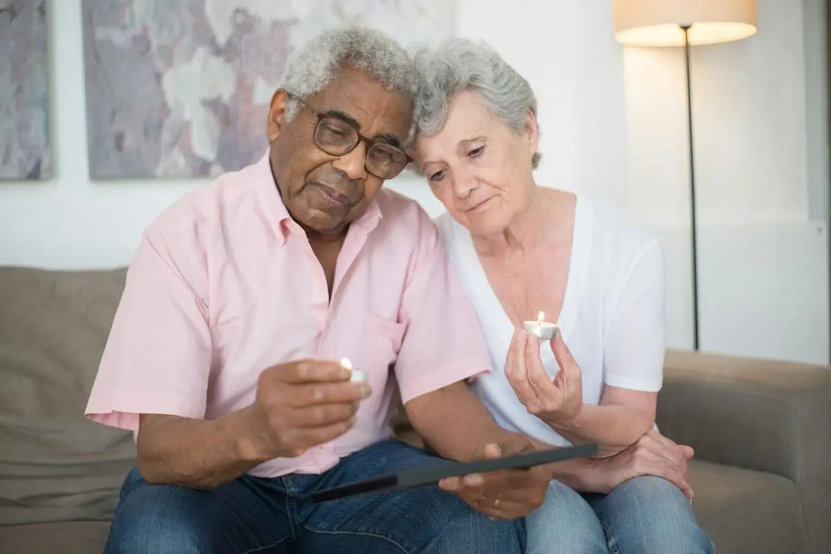 A grieving elderly couple holding candles, reminiscing over a photograph indoors.