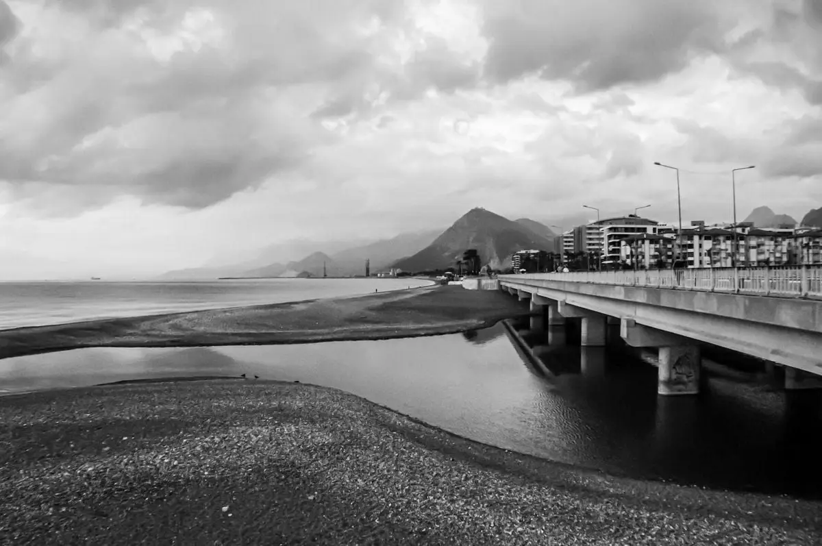 Black and white photo of a coastal landscape featuring a bridge with mountains in the background.