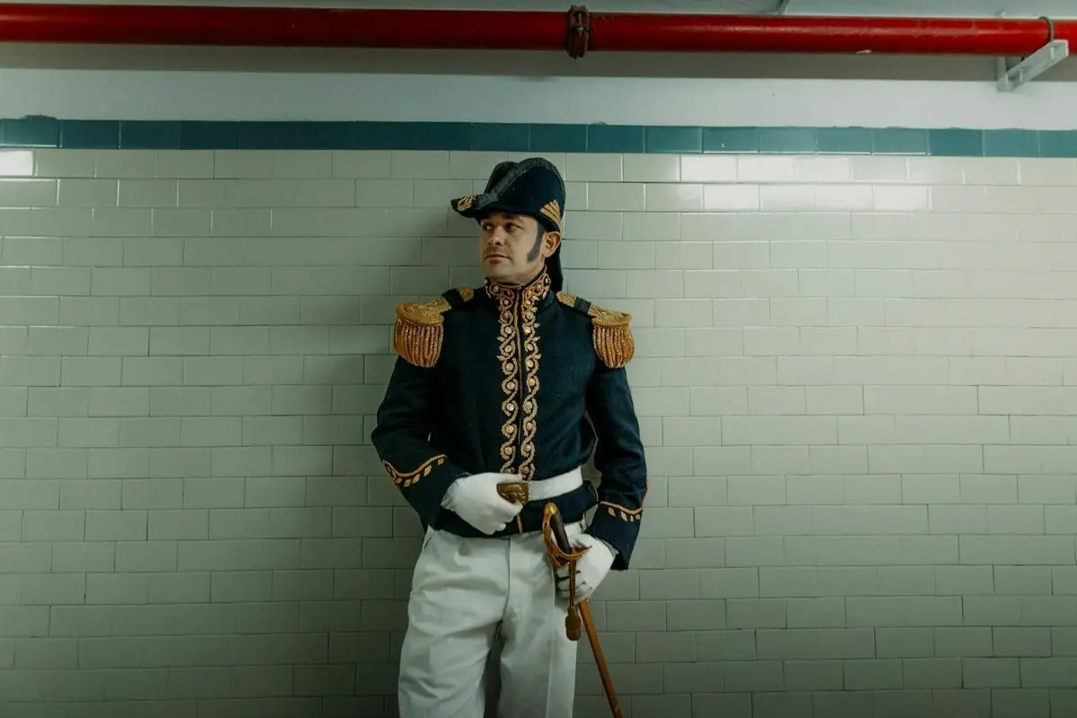 Man in a detailed historical officer uniform stands against a tiled wall.