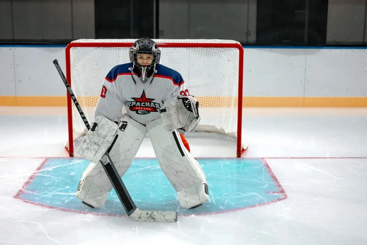 Focused ice hockey goalkeeper stands ready in front of the goal, equipped with helmet and pads on an indoor ice rink.