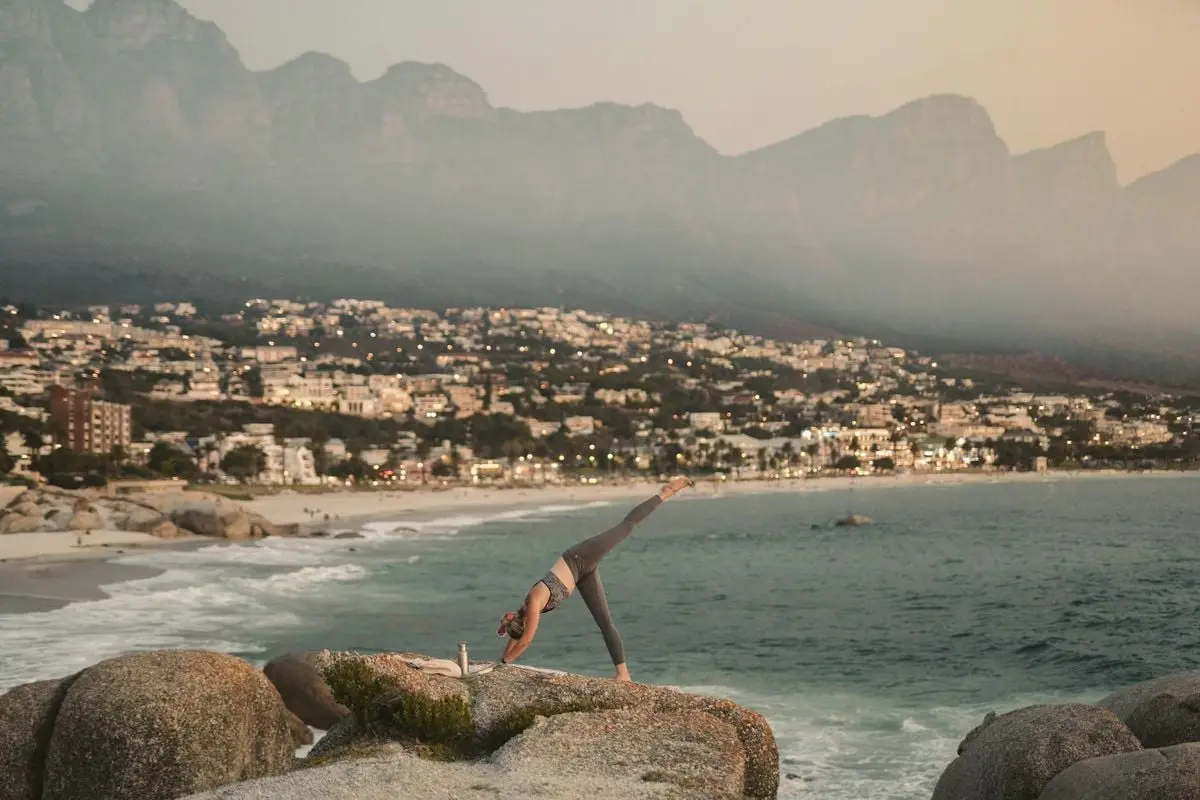A woman practices yoga on a cliff overlooking a beach in Cape Town with mountains in the background.