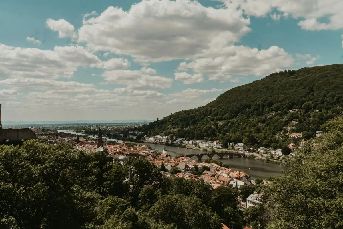 Idyllic landscape of Heidelberg with arch bridge, riverside town, and lush hills under a summer sky.