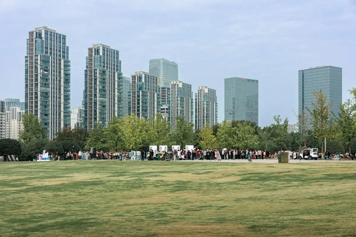 A crowd gathers in a city park with towering skyscrapers in the background under a clear sky.