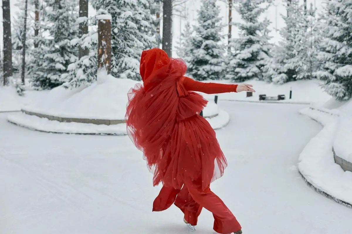 A graceful ice skater in red spins on an outdoor rink, surrounded by snow-covered trees.