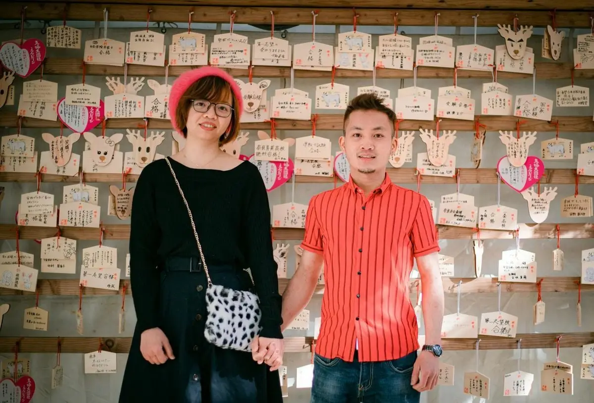 Trendy couple holding hands in front of a Japanese wish wall, showcasing love and cultural tradition.