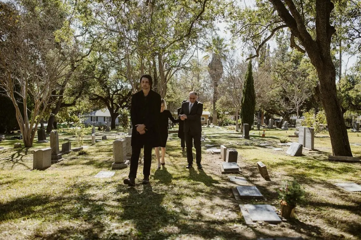 Three adults in formal attire walking through a sunlit cemetery, reflecting grief and loss.