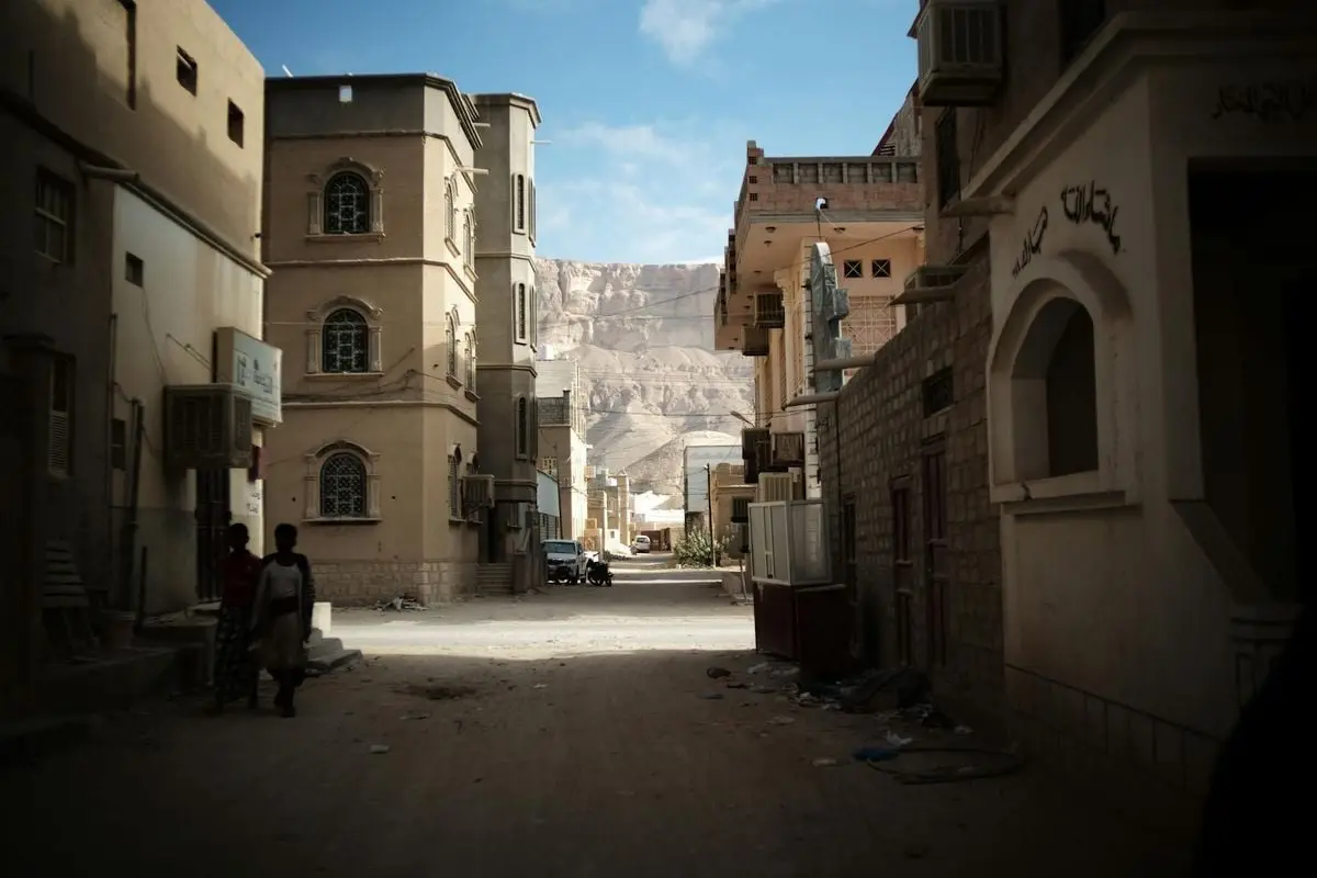 A quiet alley in a Middle Eastern town framed by traditional architecture and mountain backdrop.