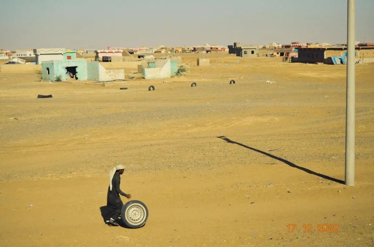A boy rolls a tire through a sunlit desert village with scattered buildings.