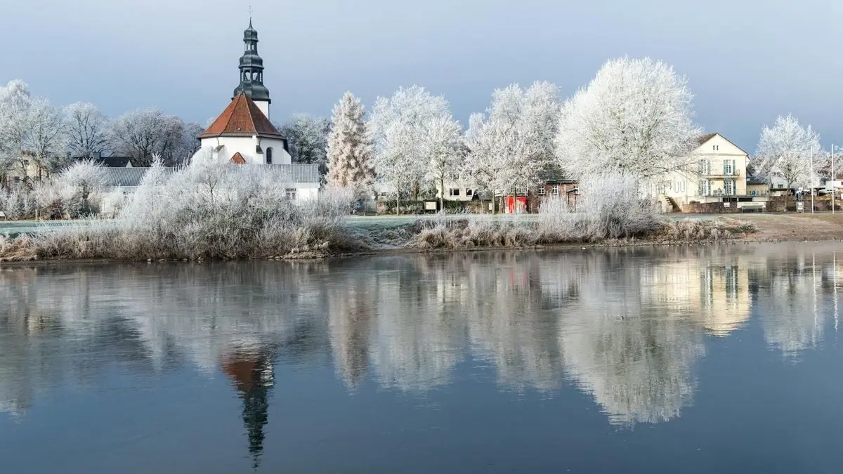 Serene winter landscape with frosty trees and church reflecting in Beverungen lake.