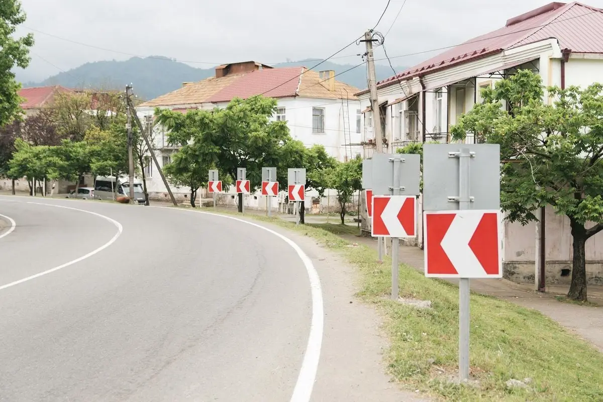 A winding road lined with arrows and trees, bordered by houses in a small town.