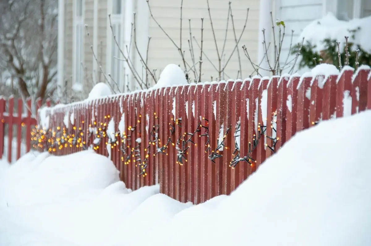 A snowy wooden fence in Finland adorned with warm festive lights reflecting a cozy winter scene.