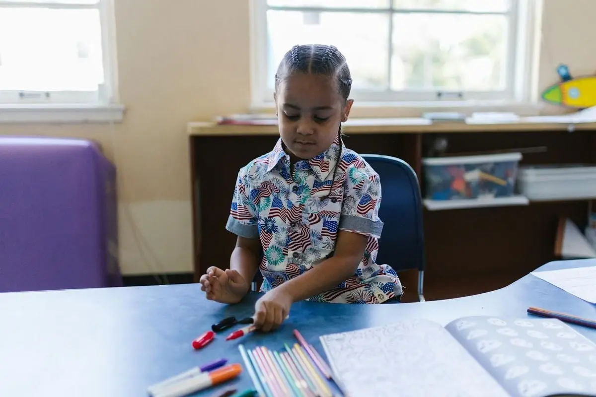 A young boy thoughtfully organizing colored markers at a school desk.