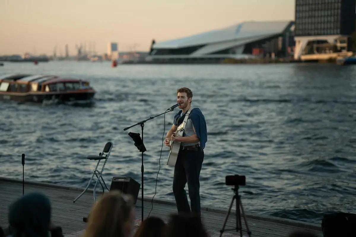 A street musician playing guitar and singing by the waterfront during sunset, with a scenic backdrop.